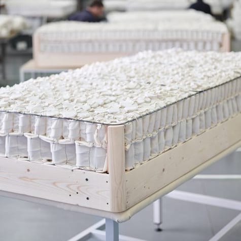 A close-up of a mattress on a wooden frame in a bright, industrial workshop setting with other mattresses and workers blurred in the background.