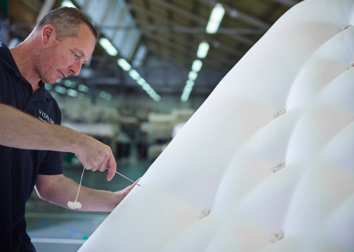 A man in a dark navy polo shirt inspects a large, white, tufted upholstered surface in a bright, modern industrial workshop or factory setting.