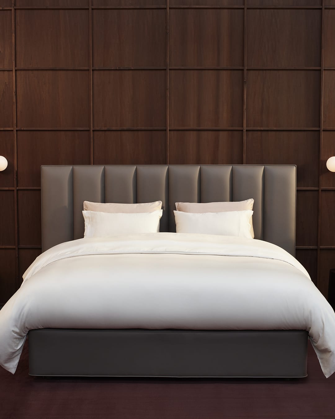 A modern bed with a padded gray upholstered headboard and matching base, dressed in white bedding, set against a dark wood paneled wall.