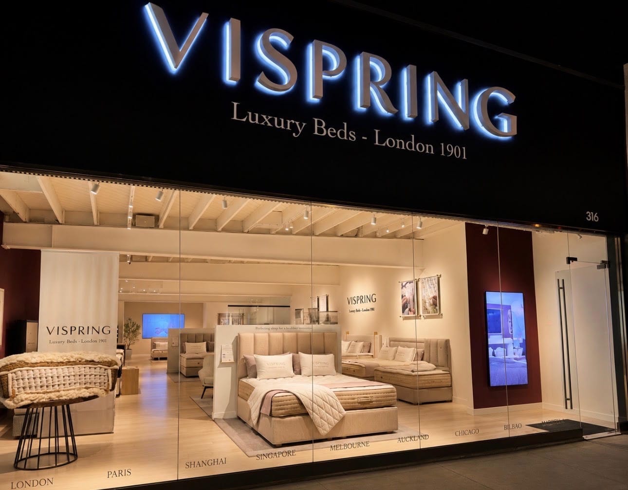 VISPRING luxury beds retail store front in Los Angeles, featuring a large illuminated black sign with blue-white lettering, a glass-enclosed showroom displaying upholstered beds and seating, and a warm beige interior.