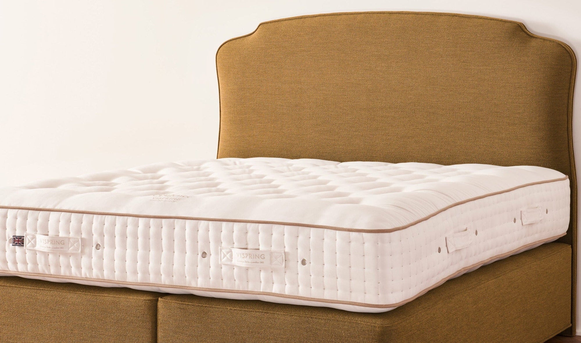 A white quilted mattress on a matching upholstered bed frame with a tall curved headboard, set against a plain off-white wall.