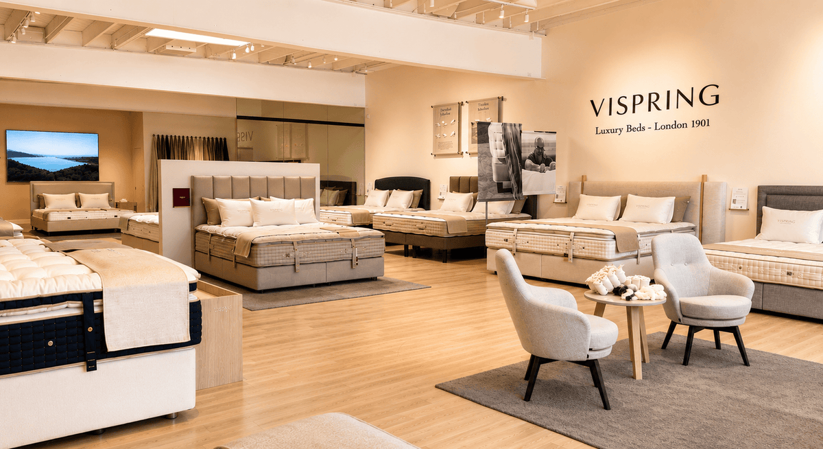 A luxury bedding showroom featuring multiple mattresses in various styles and colors, styled with neutral upholstered chairs, a small round table, and a warm wood floor. The brand name 'VISPRING' is clearly visible on the wall.