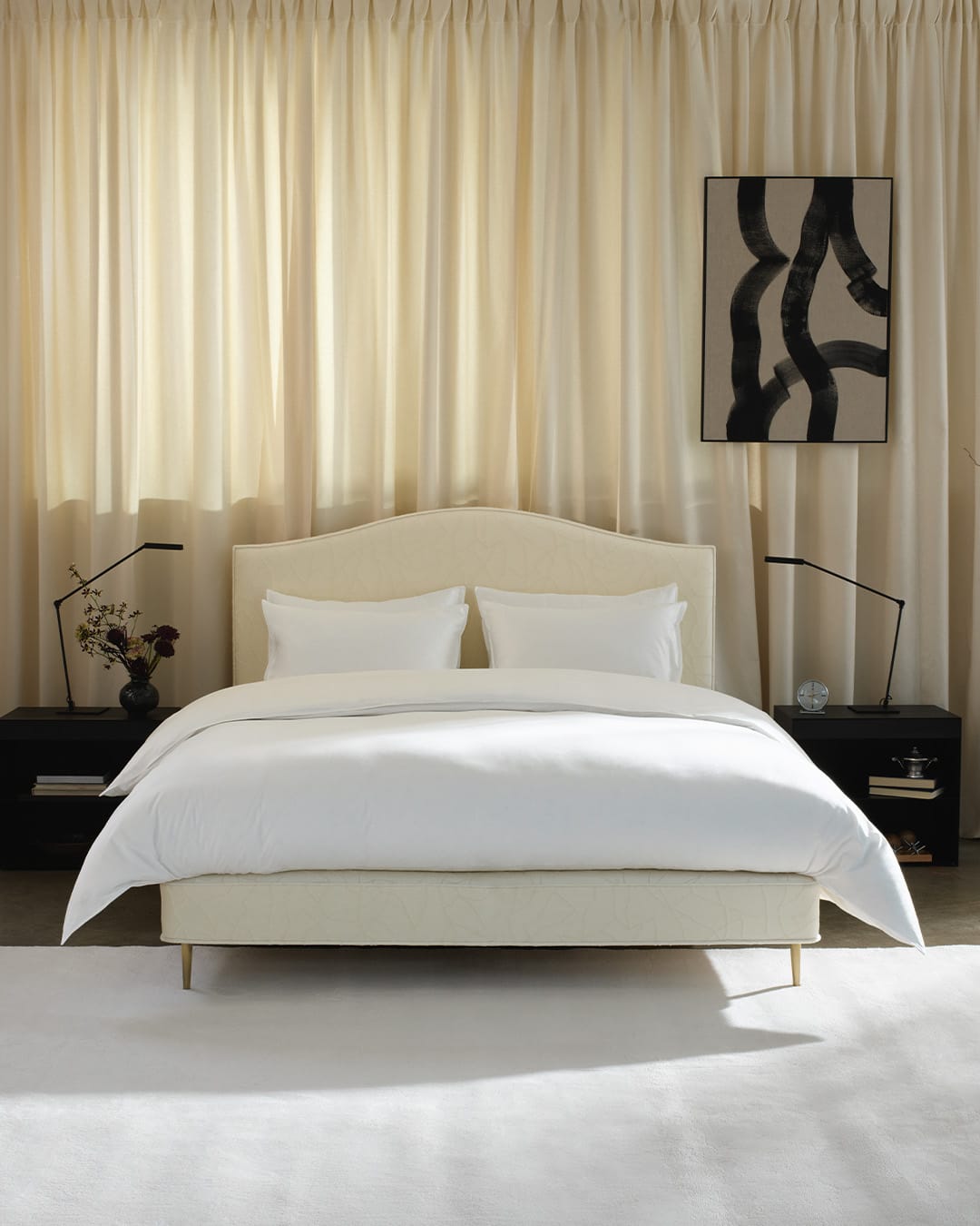 A modern upholstered bed with a curved cream headboard and matching footboard, dressed in white bedding, set against a backdrop of draped beige curtains with black nightstands and lamps on either side.