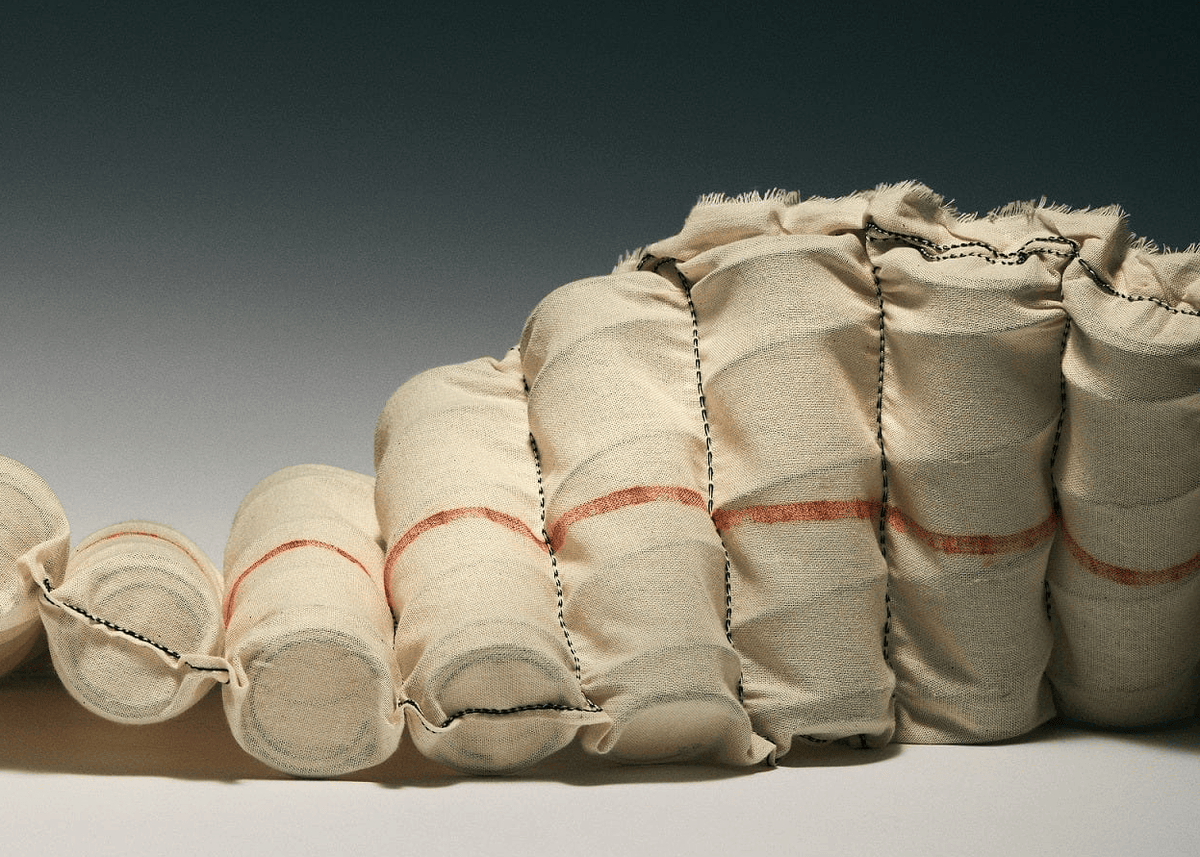 A close-up studio shot of a beige fabric-covered cylindrical support or cushion structure with visible orange-red stitched bands and dark contrast stitching, photographed against a dark-to-light gradient background.