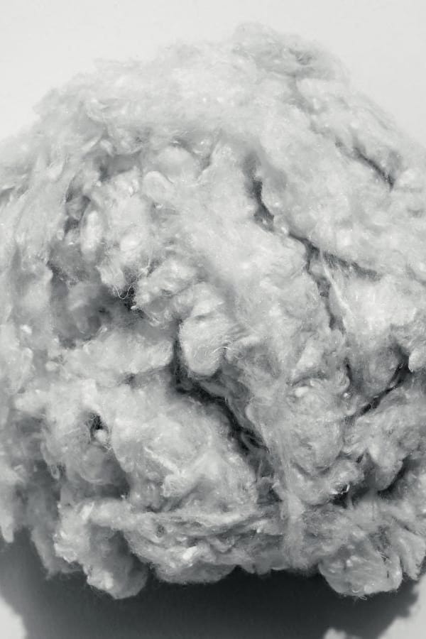A close-up of a fluffy, textured white or light gray fiber ball, likely cotton or wool, on a plain light background.