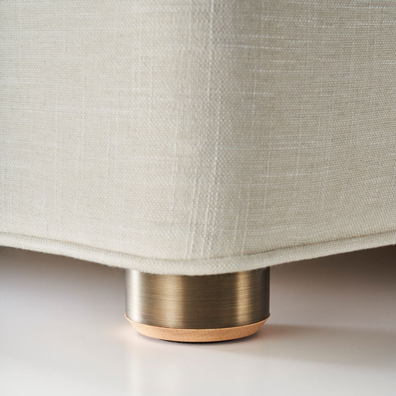 Close-up of the corner of a light beige upholstered sofa or chair with a brushed brass cylindrical leg cap and a thin wood-toned base, shown on a clean white surface.